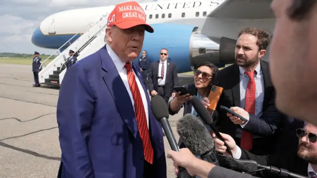 Donald Trump stands next to a white and blue aeroplane. He is wearing a white shirt, navy blazer, and a red tie with thin navy stripes on it. He is wearing a red baseball cap that says in white lettering Make America Great Again. Reporters in suits stand in front of Trump holding microphones towards him.