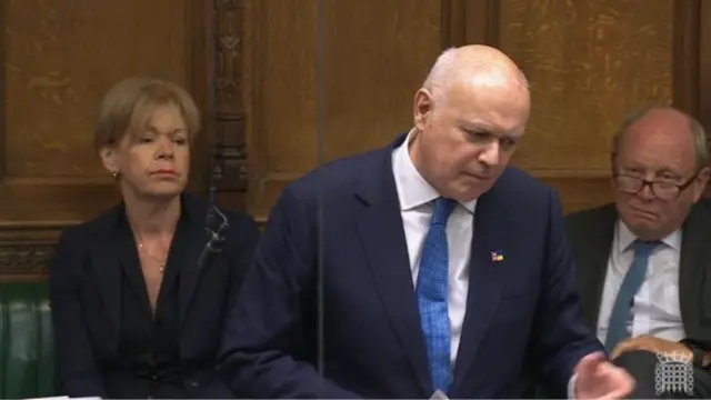 Iain Duncan Smith in a dark blue suit, white shirt and lighter blue tie, partially leaning left as he gestures while delivering a speech, two colleagues sitting on the Commons' benches behind him