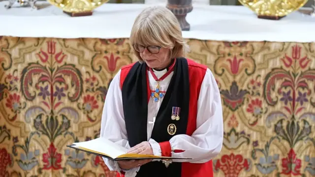 Bishop of London Sarah Mulally reads scripture off a green leather folder while wearing a white tunic with a red vest and black scarf with medals, a silver cross around her neck and a floral tapestry behind her