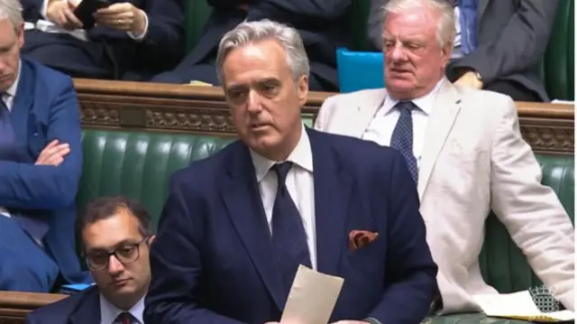 Conservative MP Mark Garnier in dark blue suit, white shirt and matching blue tie stands from the green leather benches of the Commons, colleagues sitting behind him