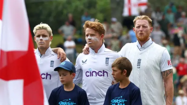 Jacob Bethell, Ollie Pope and Joe Root line up to sing the anthem in front of an England flag. There are two children as mascots between them.