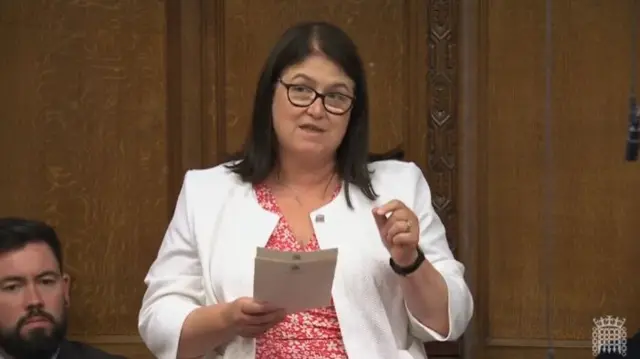 Labour MP Rachel Hopkins in a white blazer and red and white floral dress stands in the Commons to deliver speech, two sheets of paper in her right hand