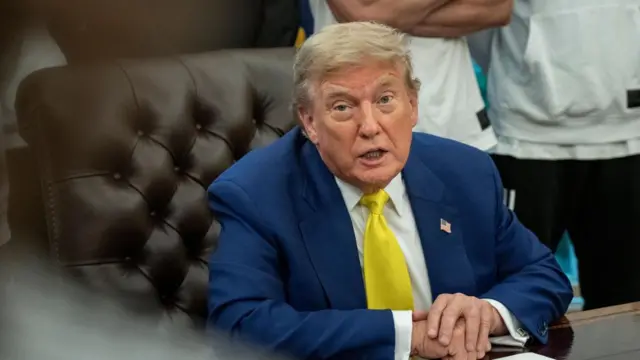 US President Donald Trump sits at a desk in the Oval Office.  He is wearing a blue suit and a yellow tie