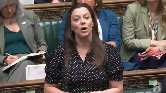 Lib Dem MP Sarah Green in black dress with small white polka dots speaks in the Commons, three colleagues sitting on the green leather benches behind her