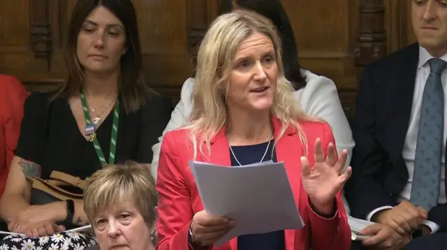 Labour MP Kim Leadbeater stands in the Commons wearing a hot pink blazer, black top with a silver chain around her neck. She's holding a small stack of papers with her right hand, other party members sitting on the benches behind her