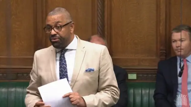 James Cleverly in tan suit, white shirt as he stands from the green leather benches of the Commons