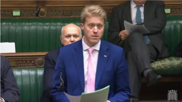 Dr Ben Spencer MP in a dark blue suit, white shirt and pink tie holding papers in his left hand as he delivers a speech in the Commons