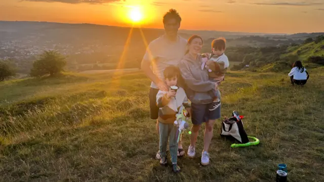 A man, woman and two children on Leckhampton Hill. The sun is rising above the country landscape in the background.