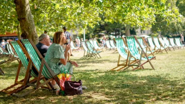 A woman in a green dress sits on a sun lounger under a tree in a park eating a cornetto. A man in a polo shirt sits to her left in a matching green-and-white chair, rows of similar chairs in front of them on the lawn