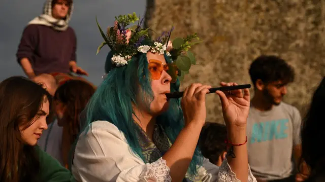 A woman with turquoise hair and a flower crown plays the fiddle as she sits on the ground next to the Stonehenge circle as the Sun rises