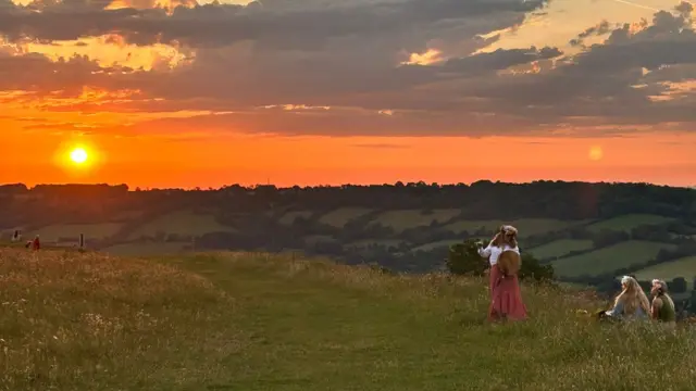 Three people on a hill near Bath. Two are sat down, with one stood up. The sun is rising over the hill in the background.