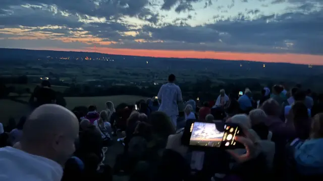 A strip of red/orange light is visible just above the horizon. In the foreground, a crowd is sitting on the hillside looking over the countryside as they wait for the sun to rise.