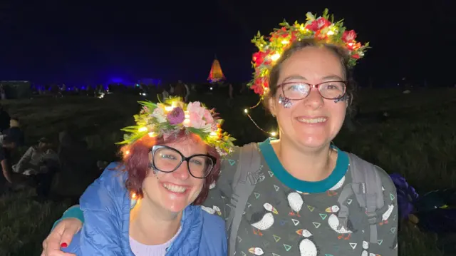 Two women huddled together smiling into the camera. Both are wearing flower crowns. Blue lights can be seen shining on the Stonehenge monument in the background.