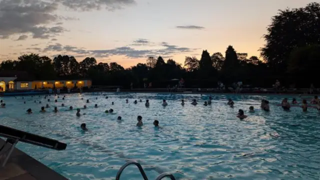 The heads of dozens of swimmers can be seen in an outdoor pool. It is dawn and the sky is beginning to light up in purple, orange and yellow hues.