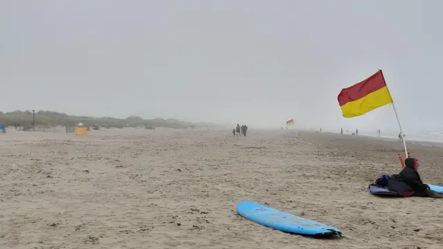 A blue surfboard sits on a beach under a very overcast sky. A person wrapped up in a coat sits next to a red and yellow flag