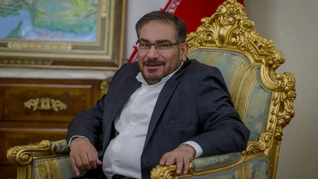 An Iranian man sits in a gilded chair while wearing a suit.