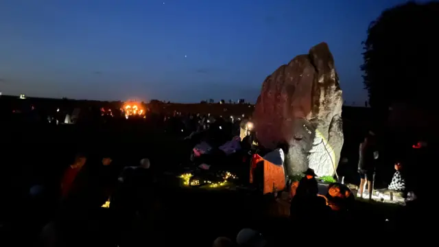 A large stone is lit up at the Avebury stone circle. A fire can be seen in the distance and people are crowded around the stone.
