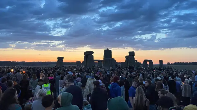 Crowds around the stones during first light at Stonehenge. Light clouds can be seen above the stones, with a strip of orange sky underneath it lit up by the rising sun - which is not yet visible. The silhouette of the stone circle can be seen behind a large crowd of people.