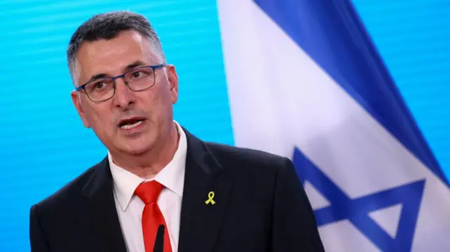 Israeli foreign minister Gideon Saar delivers speech wearing a black suit, white shirt and red tie. A yellow ribbon pin is on his left lapel, an Israeli flag behind him