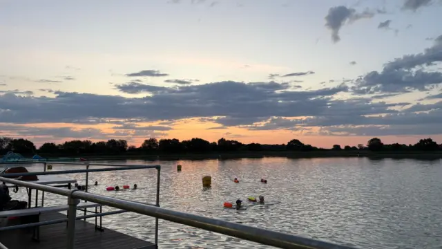 First light at West Country Waterpark, Bradley Stoke in Bristol. The sky is turning orange as the sun rises, with the colours reflecting in the water.