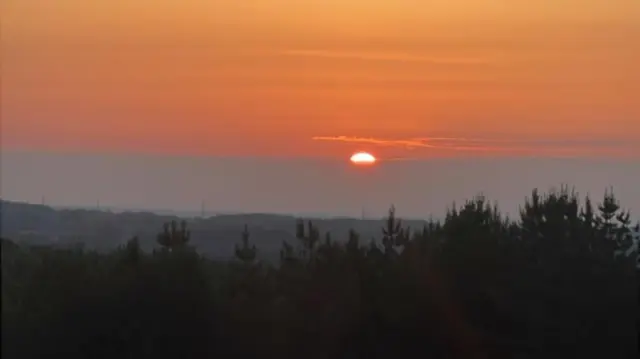 The sun peeping through the clouds and trees. The sky is a rich orange colour.