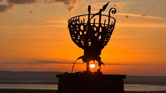 The sun rising behind a metal Welsh Dragon statue. The sea is in the distance, with the sun centre of the image.