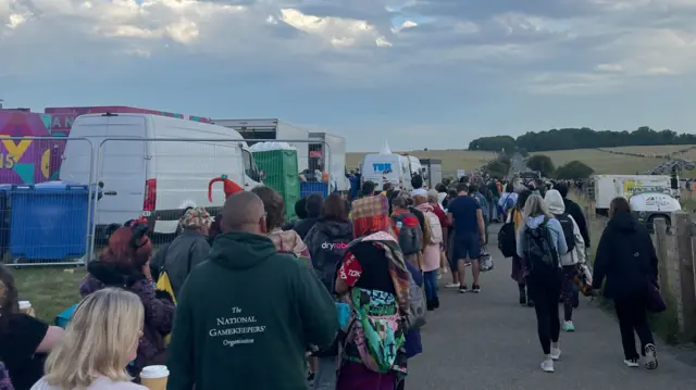 Hundreds of people leaving the Stonehenge site on foot. A queue of people can be seen in the distance, with food vans and portaloos to the left of the image.