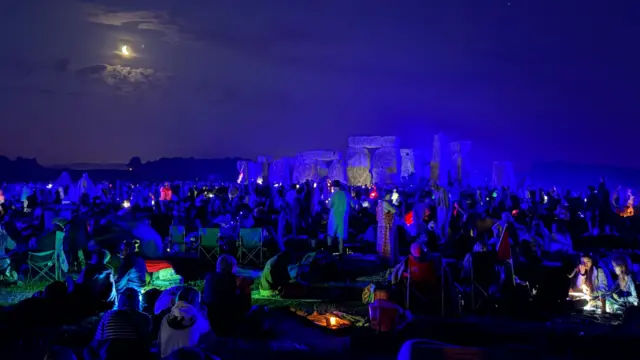 Crowds gathered around the stones of Stonehenge. The stones are lit up in blue. A bright crescent moon can be seen in the top right of the image.