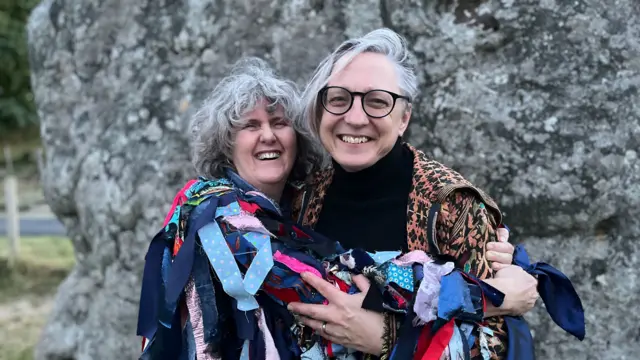 Two women embrace as they smile and pose for a photo. Grace, on the left, is wearing morris dancing attire.