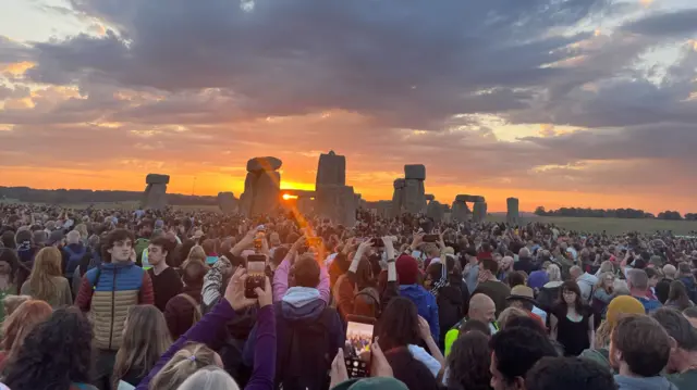 People standing looking towards the stones at Stonehenge. The sun rises above it. Lots of people use their phone camera to capture the moment.