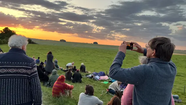 A man holds up a phone camera to the skyline, where the sun is rising. Behind is fields where people are sitting.