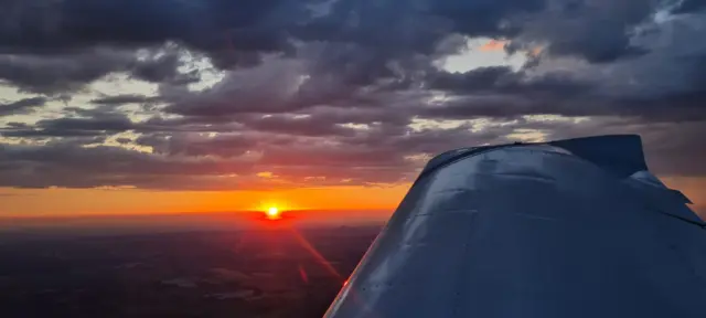 The sunrise from the air. The wing of a plane is to the right of the image, with a large amount of clouds in the sky.