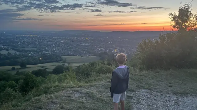 A boy stood at Leckhampton Hill looking into the distance. It is dawn with a hazy, orange light filling the sky.