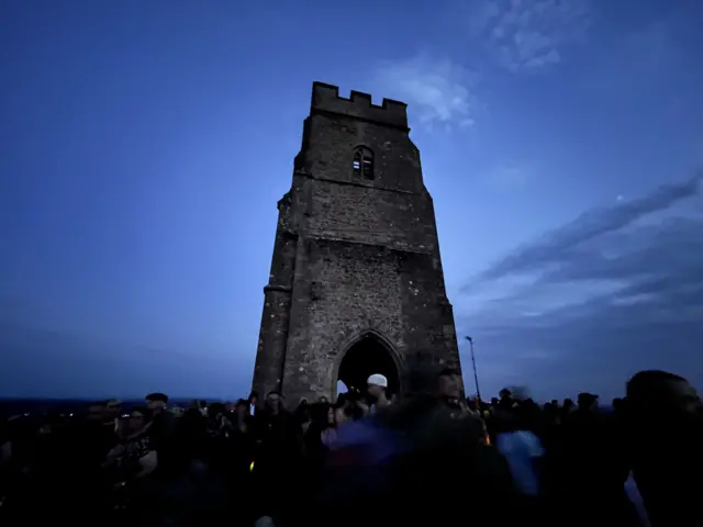 A crowd is gathered underneath a tall church tower which sits on top of Glastonbury Tor. It is dawn and the first pieces of sunlight have lit up the sky in a light blue colour.