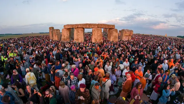 Large crowds at Stonehenge wait for the sun to rise, early light reflecs on the stones