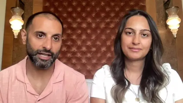 A man wearing a pink shirt and woman in a white top speak.