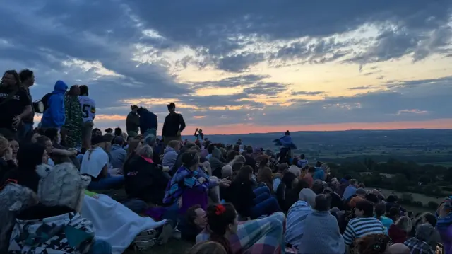 There are hundreds of people sitting on the hillside at Glastonbury Tor. Many have blankets wrapped around them as they await the sunrise. The first signs of light are visible.