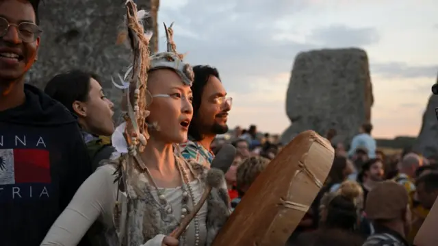 A woman with a skull headdress plays a large tambourine as she watches the Sun rise in Stonehenge, a large crowd around her and three of the stones visible in the background
