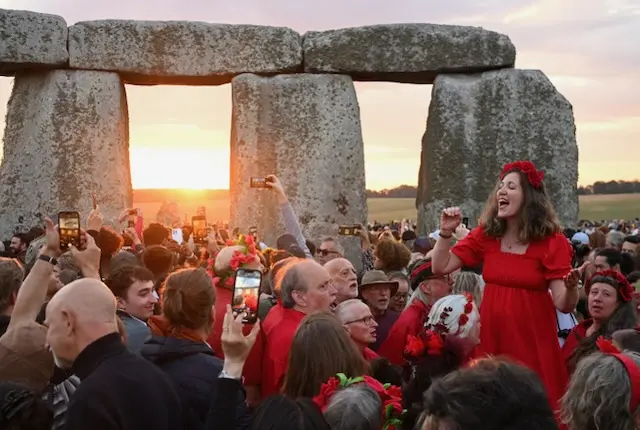 A woman in a red dress and flower crown is stood, speaking to the crowd near to the base of the Stonehenge monument. The sun can be seen shining through some of the stones.