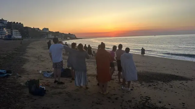 A crowd of people gathering on a sandy beach. The sun is rising over the sea in the distance.