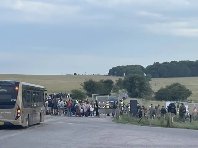 Queues of people leaving the Stonehenge site. There is a bus parked to one side.