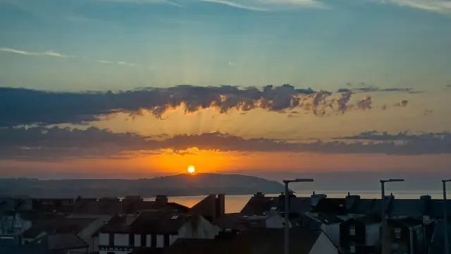 The sun rising over a hill and the sea. The rooves of buildings take up the bottom of the image.