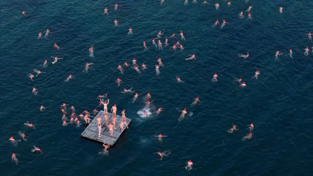 A drone captures dozens of nude swimmers, all wearing the same red caps, swimming around a ponton in a deep blue ocean. About six people are stood on the pontoon and look as though they are about to jump into the water.