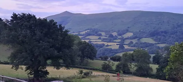 Sugar loaf mountain at first light. The sky is a light purple colour and the peak of the mountain can be seen in the distance, with hilly fields in the foreground.