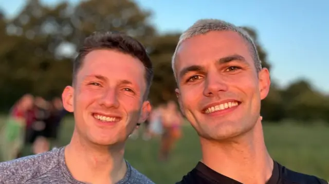 Two men smiling into the camera in a selfie photo. Their faces are lit up in golden hues from the sunrise.