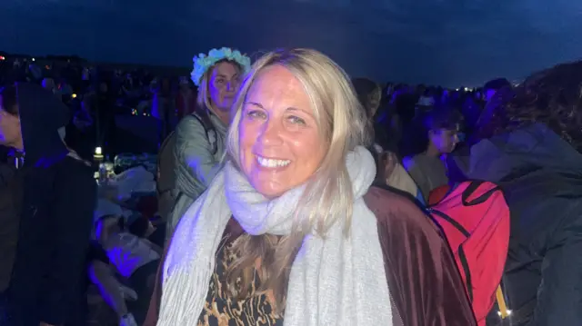 A blonde woman stands in a crowd and smiles for a photo. She is wrapped up in a grey scarf and brown coat. A crowd can be seen in the background.