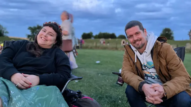 Matthew and Sabina sat on camping chairs at Avebury. They are both smiling at the camera. It is dawn, with a low light surrounding them.