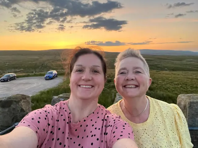 Laura and Leslie smiling into the camera for a selfie. They are in the countryside and the sunrise has created a beautiful orange glow over grassy fields in the background.