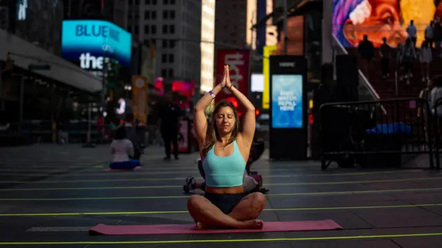 A woman sits on a yoga mat in Times Square. She is sat with her legs crossed and has her arms stretching above her head with her hands in a prayer position. Some of Time Square's billboards can be seen in the background.