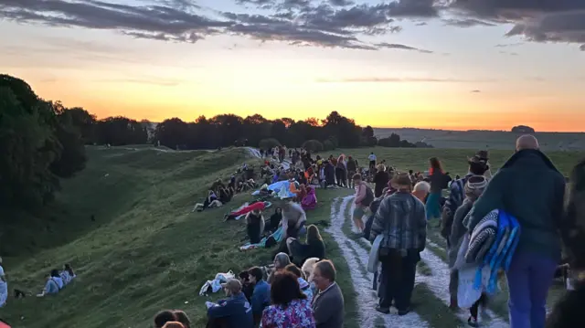 Dozens of people sit on a steep hill alongside a footpath, with more people walking along with picnic blankets in hand. The sun is beginning to rise, with the sky lit up in yellow and orange hues.